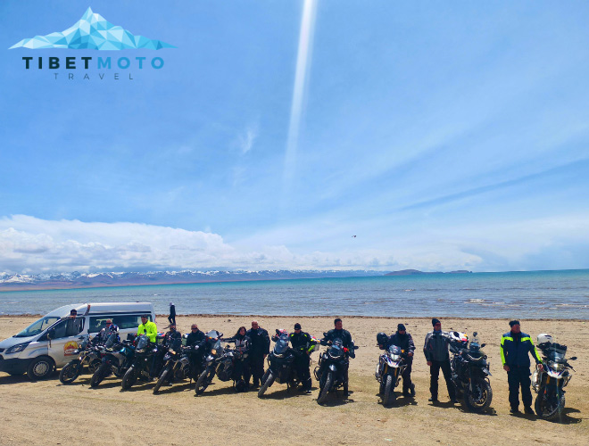 Gruppenfoto vor einem See in Tibet Mehrere Motorradfahrer posieren für ein Gruppenfoto am Ufer eines Sees in Tibet. Der See erstreckt sich bis zum Horizont, dahinter erheben sich schneebedeckte Berge.